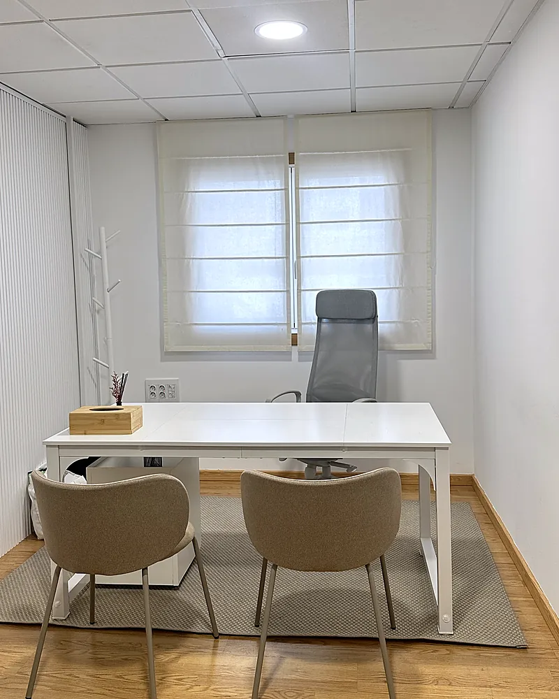 Consulta con mesa blanca, dos sillas tapizadas y ventana con estor